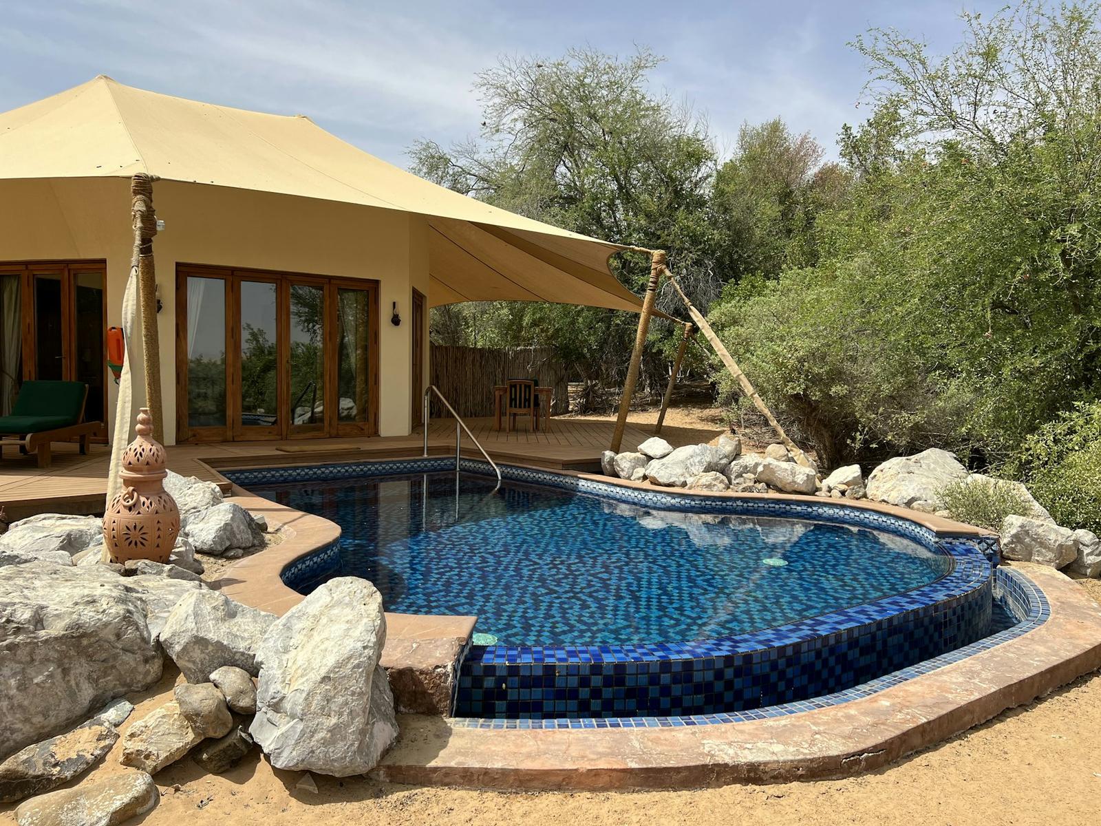 A freeform pool in blue mosaic tile, edged with boulders and desert sand